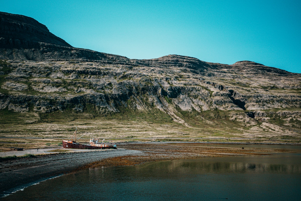 A shipwreck rests on a rocky Icelandic shore beneath dramatic cliffs and mountains