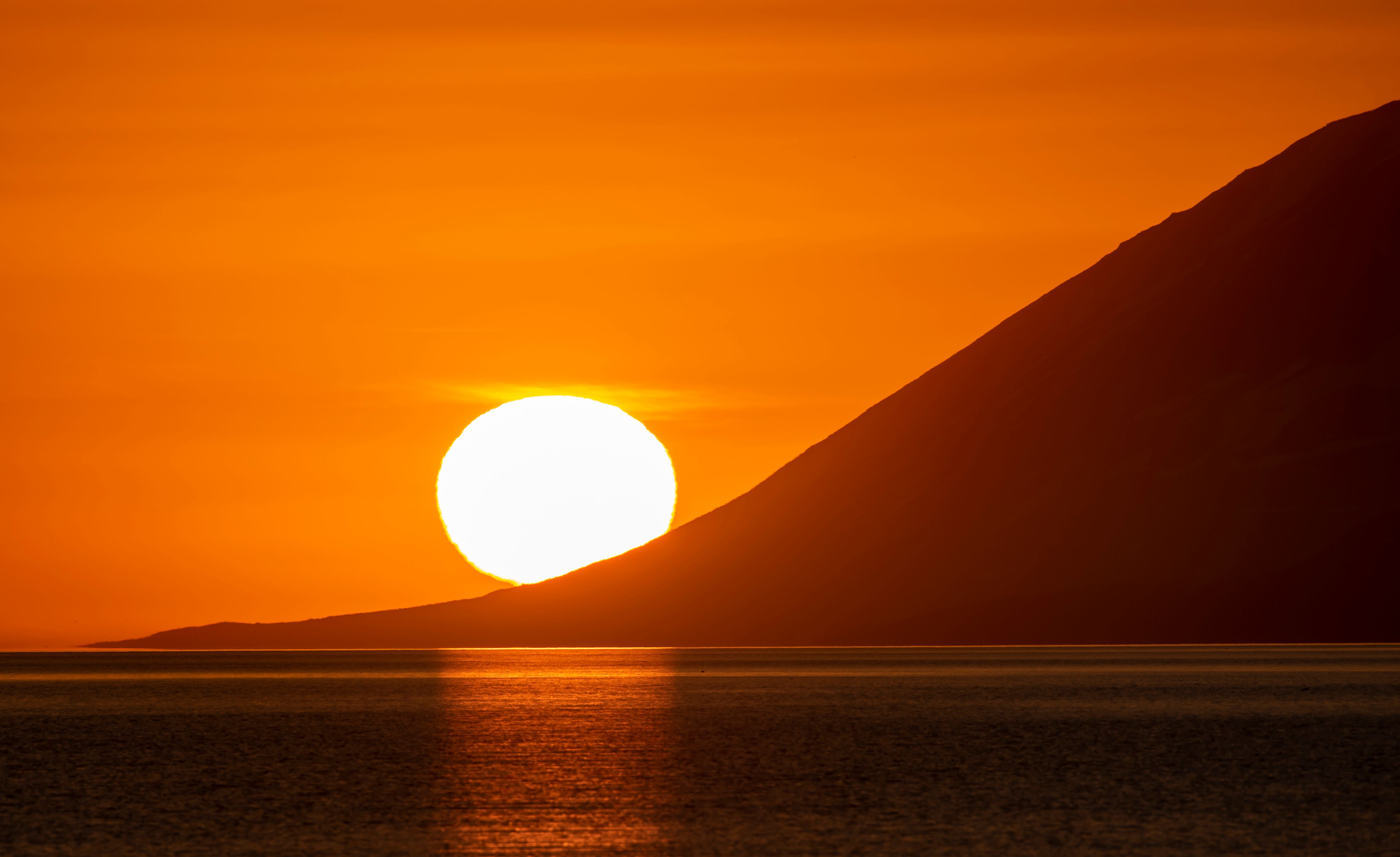 The midnight sun in Iceland — a brilliant white sun low on the horizon above a mountain silhouette and calm ocean, deep orange sky at midnight in June