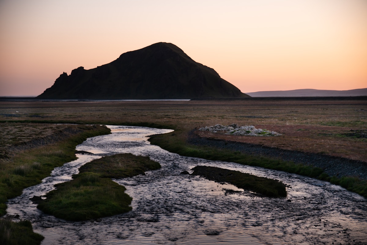 Stream winding through a flat volcanic plain toward a dark mountain silhouette under a glowing sunset sky, Iceland midnight sun
