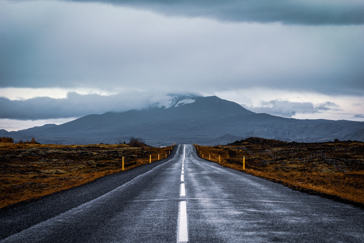 Straight paved road heading toward a snow-capped volcanic mountain under dark clouds, Iceland