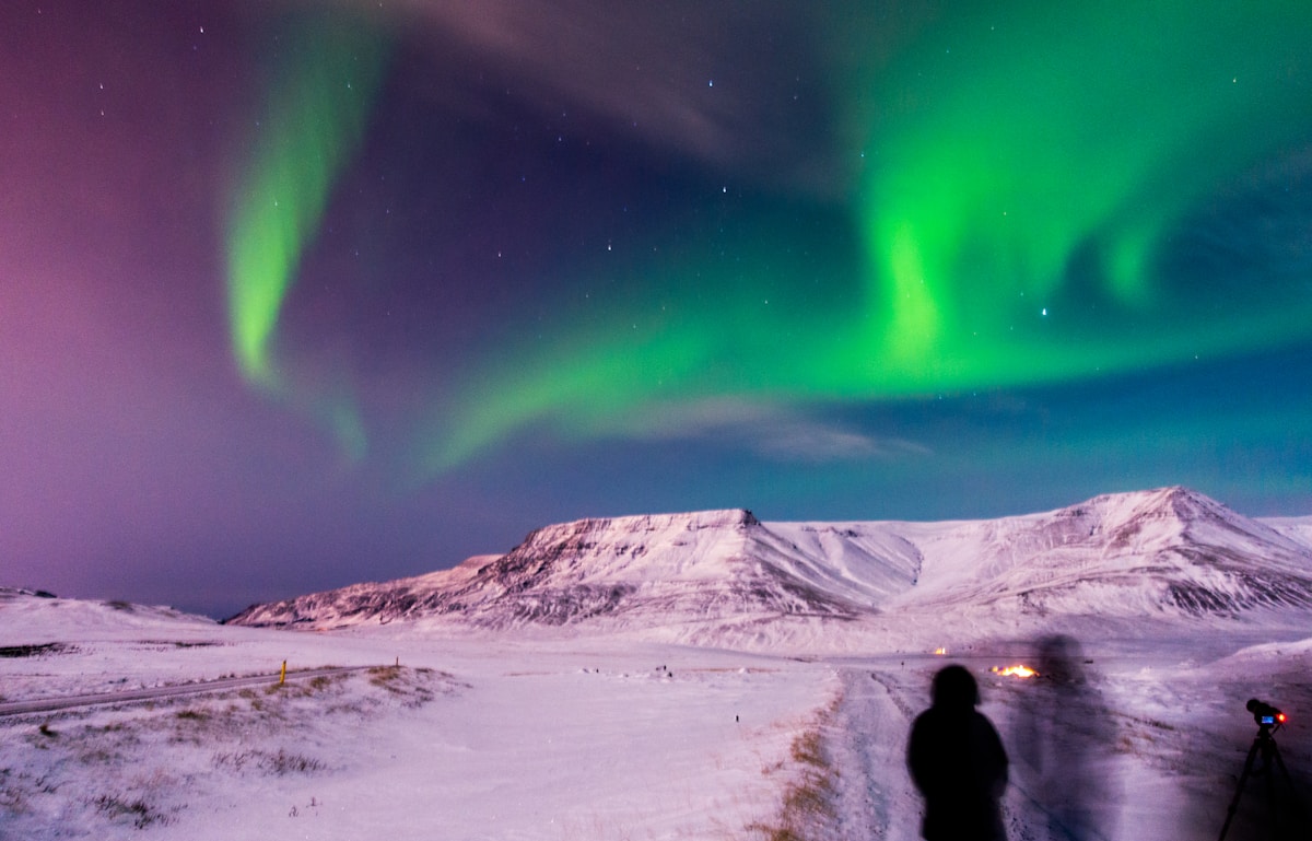 Person standing under vivid green aurora borealis over a snow-covered Iceland landscape at night