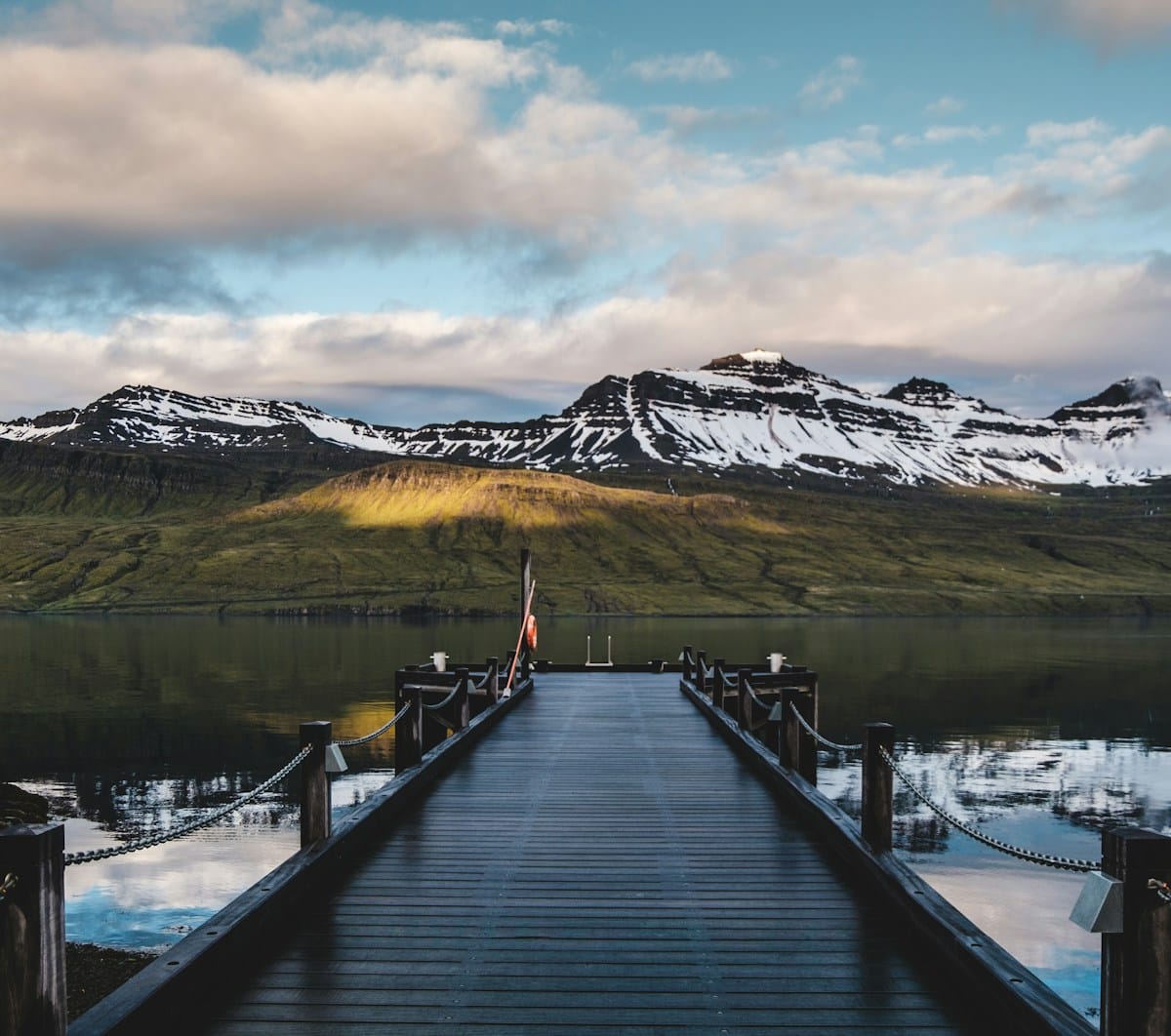 Wooden pier leading into a calm fjord with snow-capped mountains and dramatic clouds