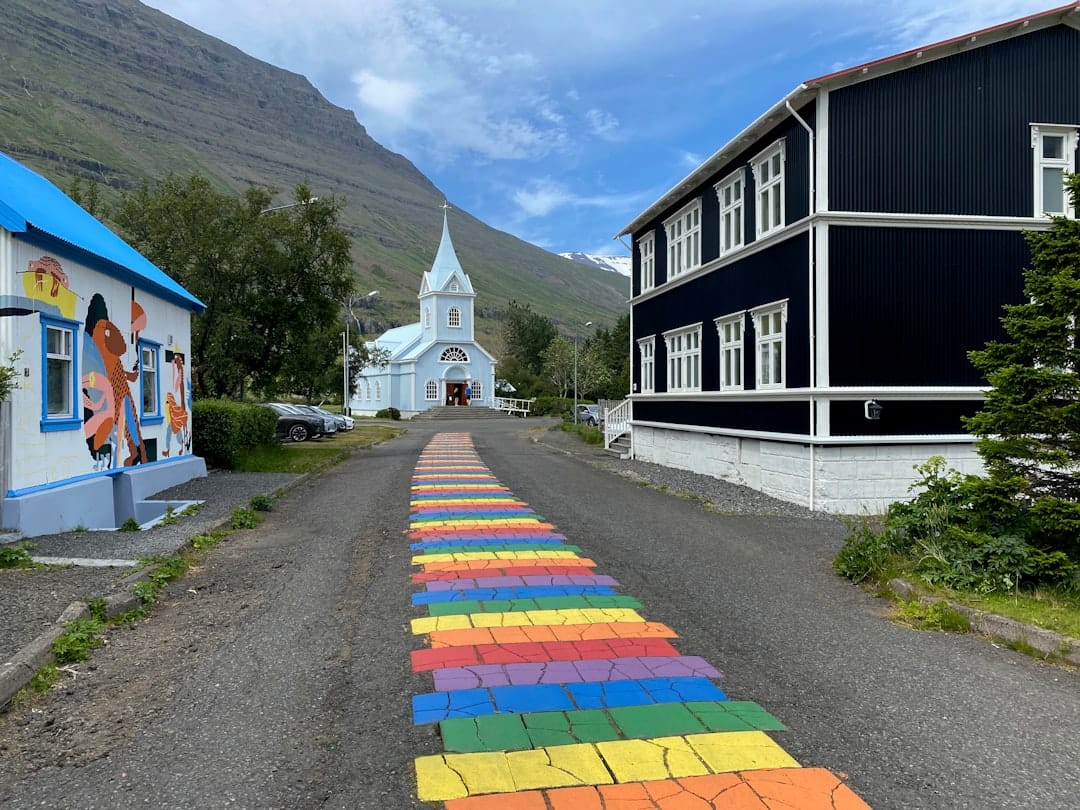 Rainbow painted street in Seyðisfjörður leading to the blue Tvísöngur church